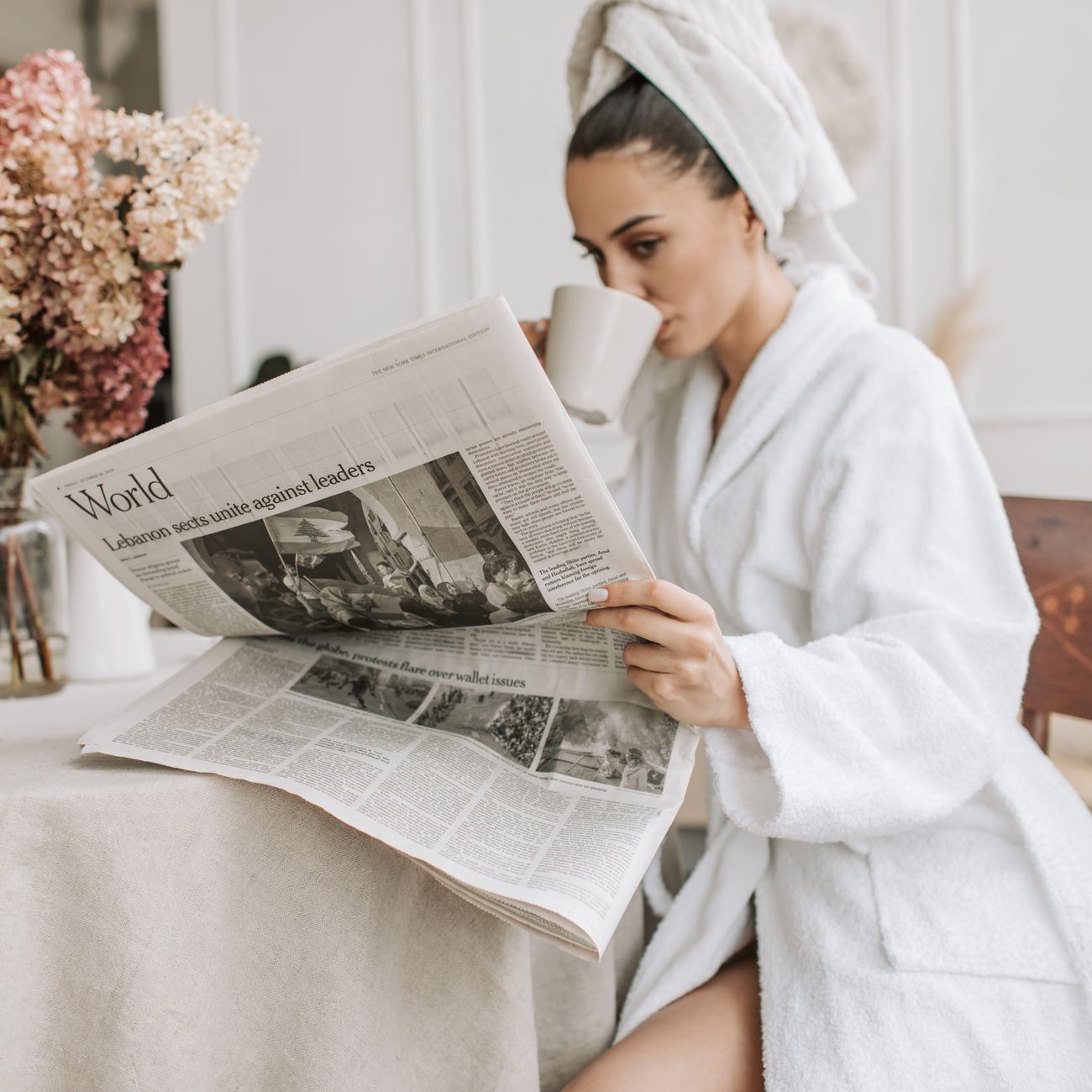 woman in white robe drinking a mug of hot drink while reading a newspaper